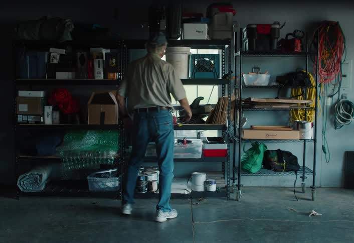 A man wearing a baseball cap and jeans organizes cluttered shelves in his garage, looking for tools and supplies