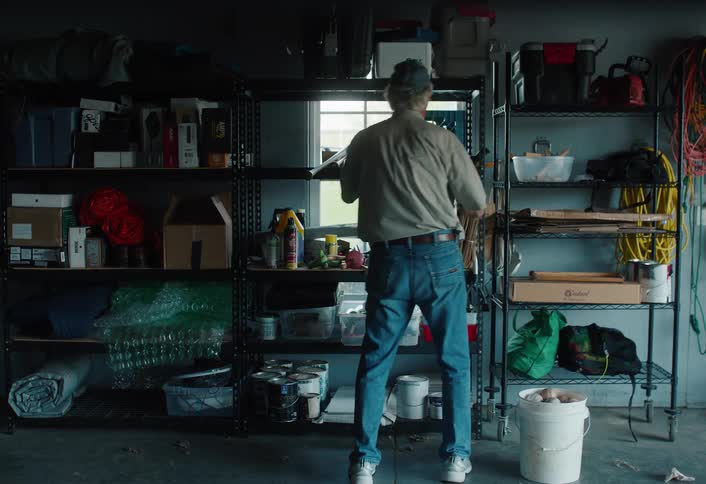 An older man wearing a cap and jeans organizes items on storage shelves in his garage