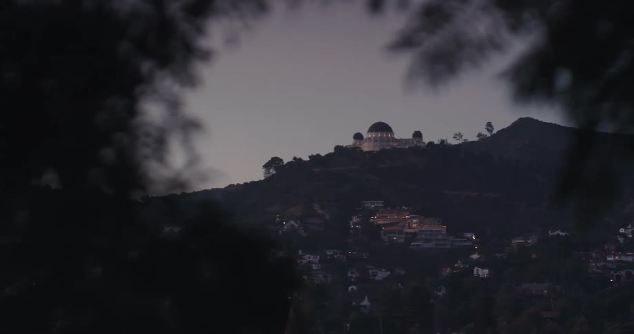 The Griffith Observatory is slowly revealed on a hilltop over Los Angeles as evening approaches, framed by dark foliage in the foreground