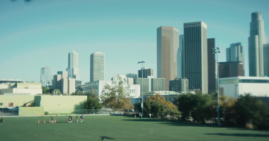 A group of people train on a green sports field in a park with the Downtown Los Angeles skyline in the background on a clear sunny day