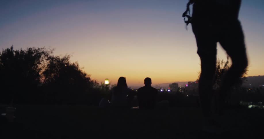 A silhouetted couple enjoys a tranquil sunset over the city from a grassy park as others walk by in slow motion