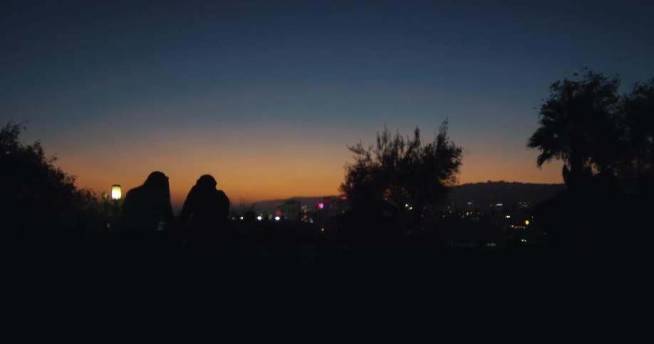 A silhouetted couple sits close together on a hill watching the beautiful sunset glow fade over the distant city lights