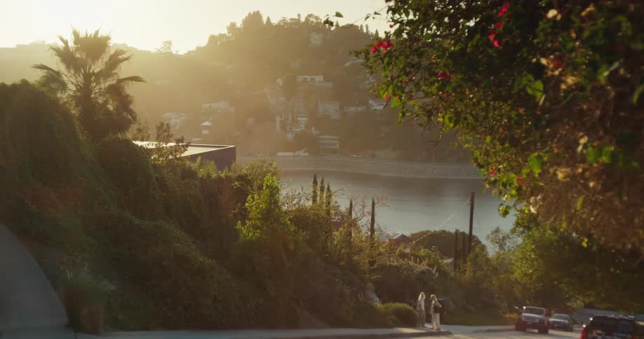 A serene, wide shot of two women walking down a lush, green hillside road overlooking a reservoir during a hazy golden hour sunset