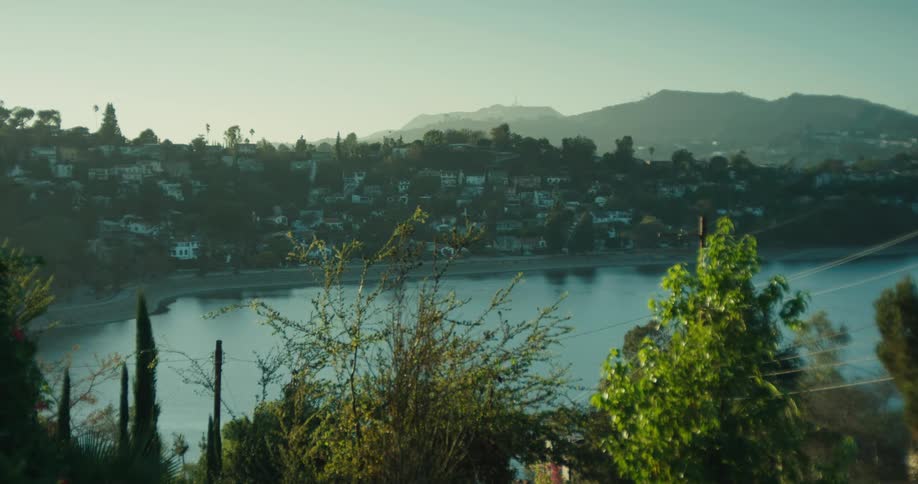 A serene, static view of the Silver Lake Reservoir with the iconic Hollywood sign visible in the distant hazy hills of Los Angeles