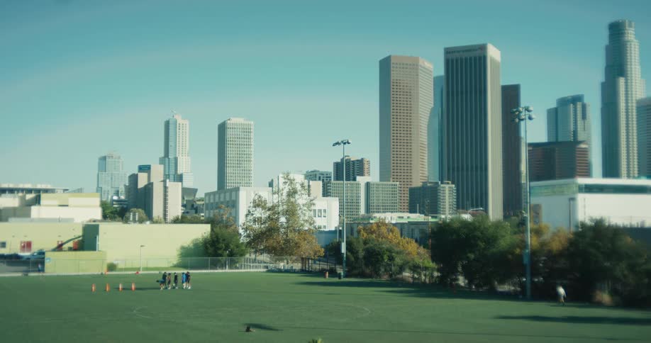 A slow rack focus reveals a group of athletes practicing on a sports field with the Los Angeles downtown skyline behind them