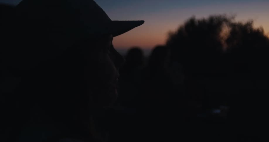 A silhouetted person in a cap takes a drink and smokes while watching the last light of sunset