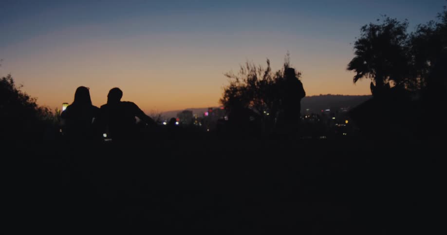 A rack focus reveals silhouettes of people watching the colorful twilight sky and city lights from a park overlook