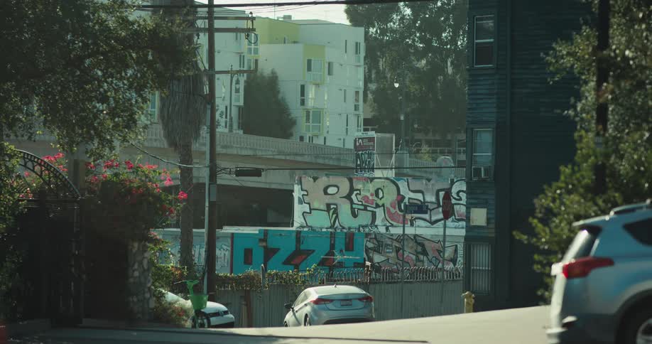 Cars drive up a hazy city street past a graffiti-covered wall, with a modern apartment building and highway overpass in the background