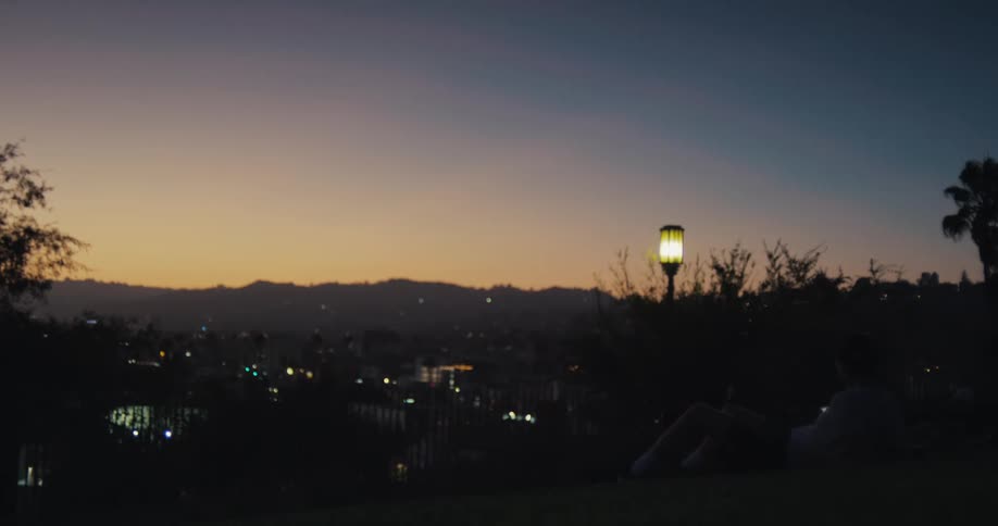 A person relaxes on a grassy hill, watching the city lights come on at dusk under a colorful sunset sky