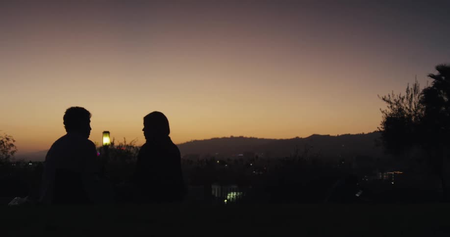 A silhouetted couple sits on a grassy hill at dusk, talking and enjoying the scenic view of the city lights below