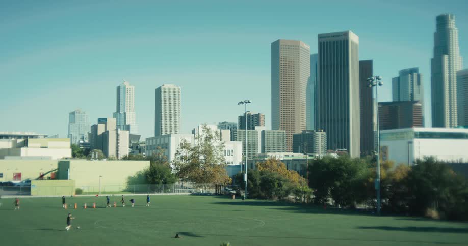 Kids practice soccer on a green field in an urban park with the Downtown Los Angeles skyline in the background