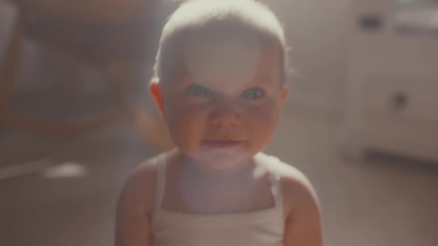 An adorable baby sits in a soft, hazy sunbeam and happily raises its arms into the air in slow motion