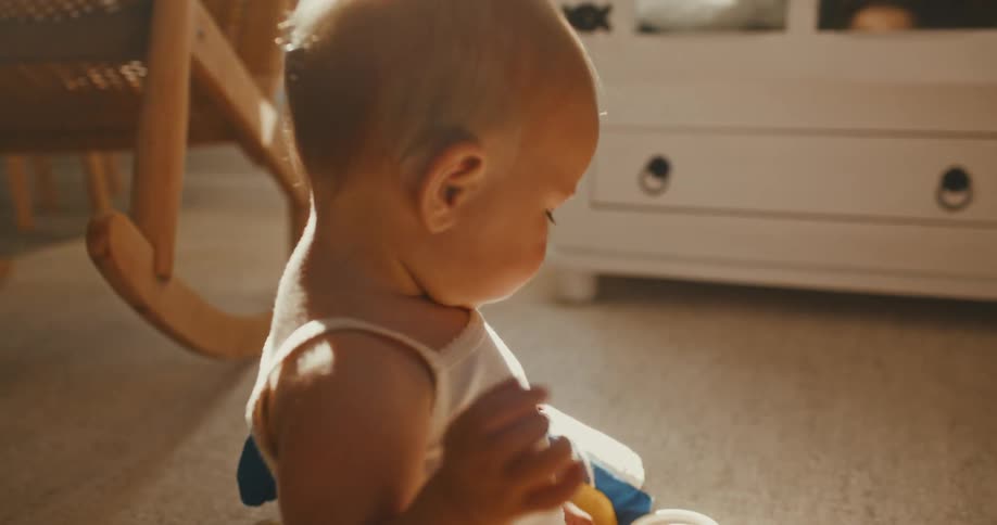 A curious baby sits on the carpet and plays with toys in a warm, sun-drenched room in slow motion