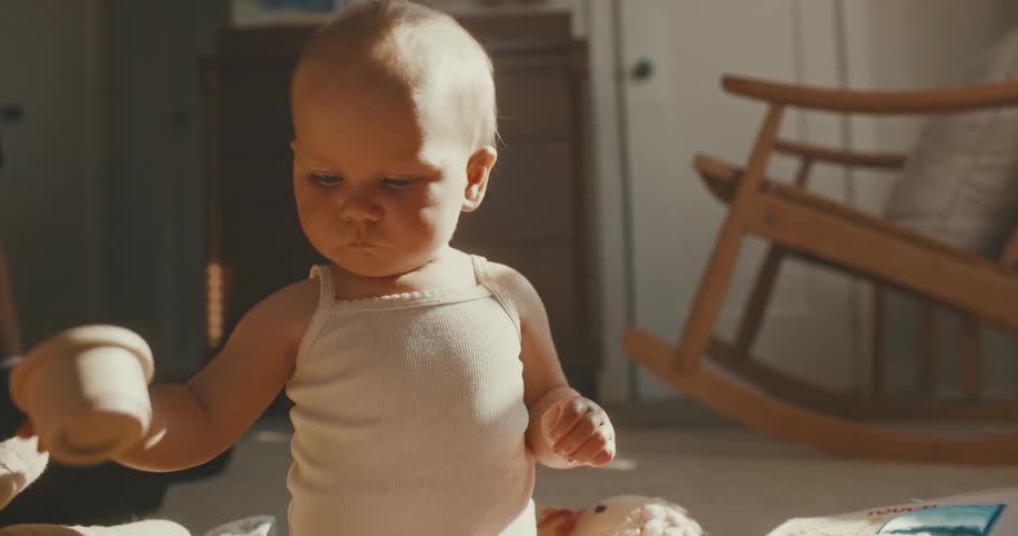 A focused baby in a white tank top plays with stacking cup toys on the floor of a sunlit room
