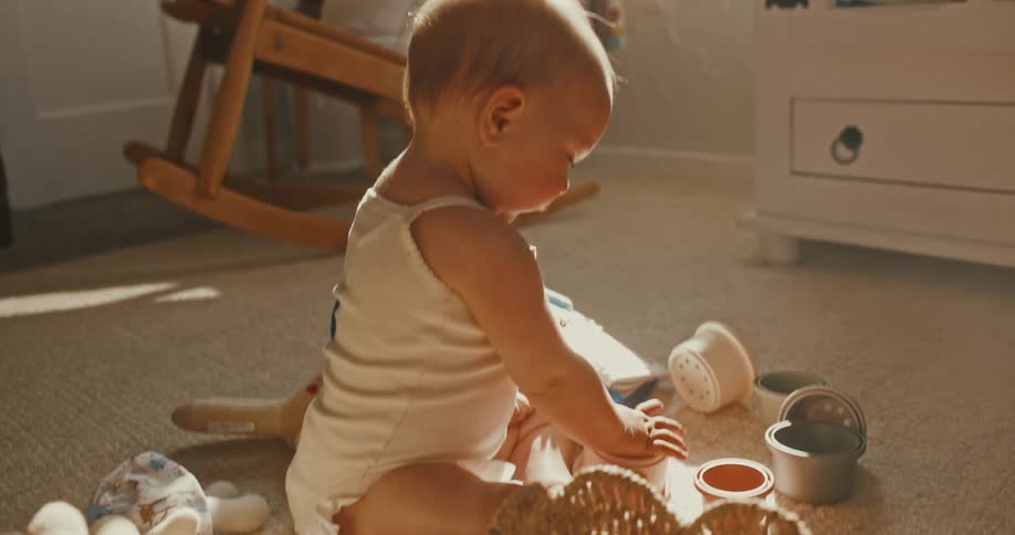 An adorable baby happily plays with stacking cups on the carpeted floor of a warm, sun-drenched nursery