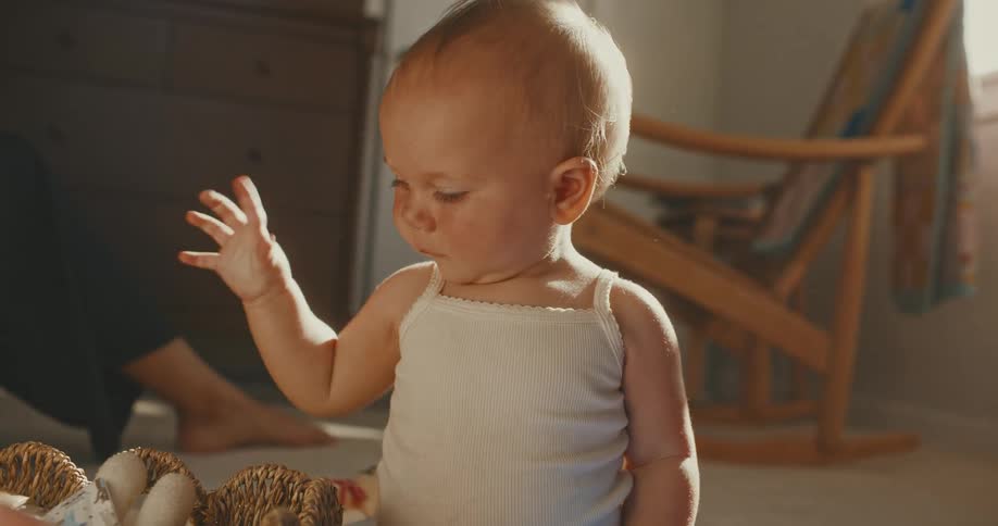 An adorable smiling baby sits on the floor in a sunlit room and playfully shakes a stuffed bunny rabbit toy