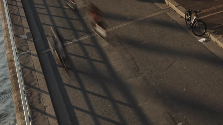 A high-angle time-lapse shows the motion-blurred movement of cyclists and runners along a riverside path, crossed by long shadows from a bridge railing