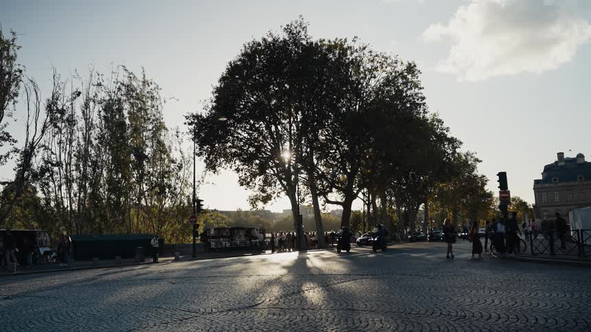 Motorcyclists and pedestrians travel down a sun-drenched cobblestone street in Paris as the sun sets behind silhouetted trees