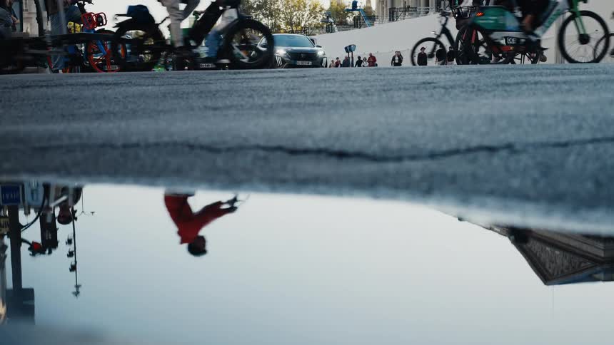 Reflections of cyclists and cars dance in a street puddle, capturing the vibrant energy of a busy city from a unique low-angle perspective