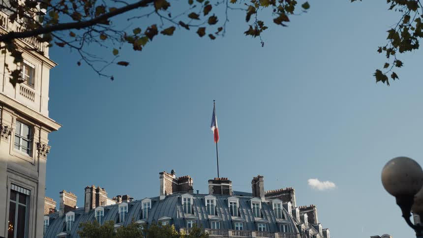 A French flag flutters atop a classic Parisian building against a clear blue sky, framed by autumn leaves