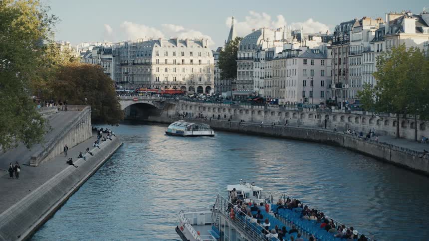 Tour boats cruise down the Seine River in Paris as people relax on the banks on a sunny day
