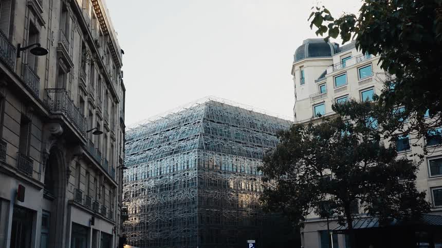 A slow tilt-down shot showcases a striking modern building with a complex geometric facade nestled among traditional European buildings on a city street
