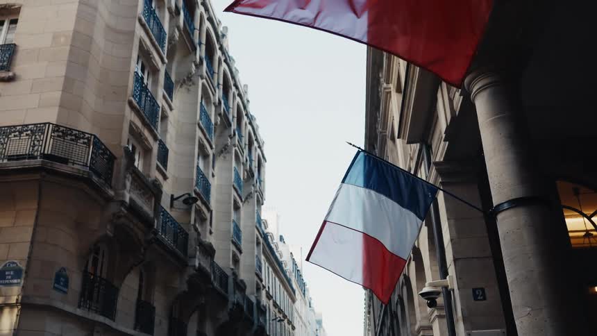 A low-angle shot shows the French flag hanging from a classic stone building on a city street in Paris