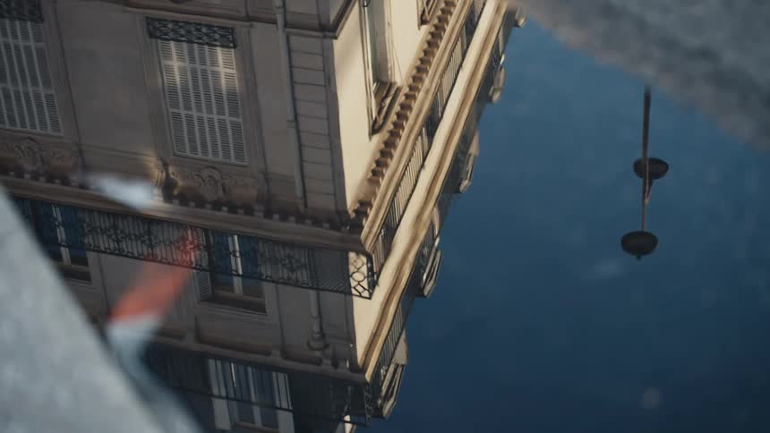 A classic Parisian building with balconies and a street lamp are reflected in a still puddle after the rain