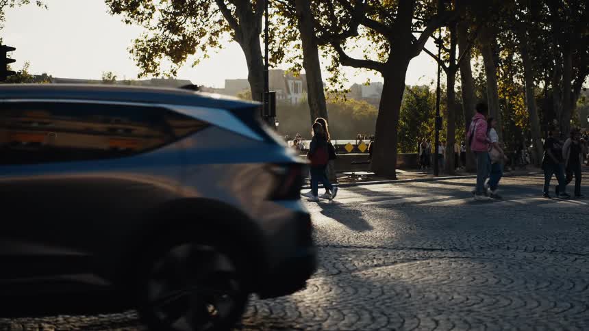 Pedestrians and cars move in slow motion across a sunlit cobblestone street in a European city during golden hour