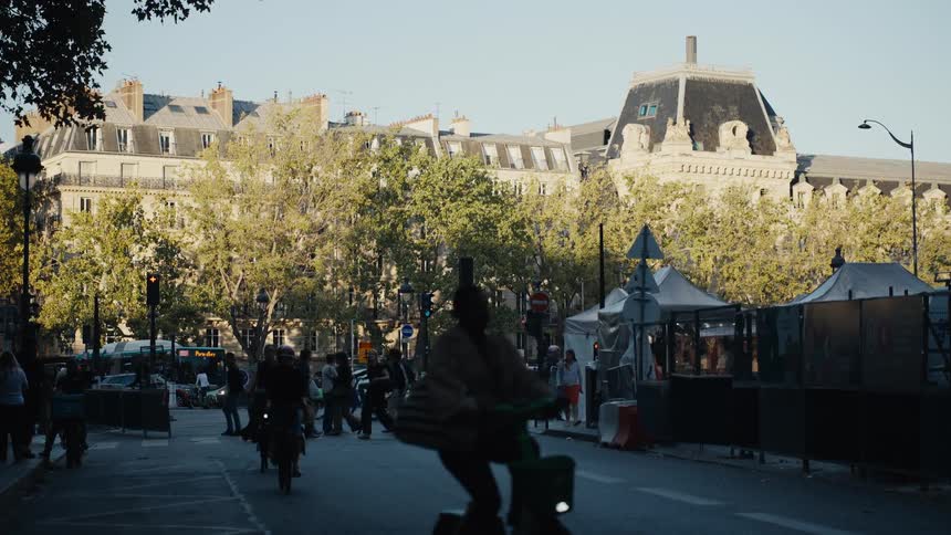 A slow-motion view captures the bustling energy of a Parisian street corner as pedestrians cross and cyclists commute during the warm golden hour light