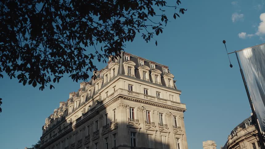 A slow pan reveals a classic Parisian building with a mansard roof under a bright blue sky, framed by silhouetted tree leaves
