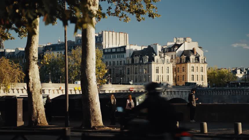 Classic Parisian architecture serves as a backdrop for a busy street with traffic and pedestrians strolling along the river on a sunny day