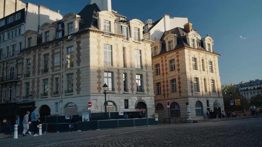 Golden hour sunlight illuminates a classic Parisian street corner as people on bicycles and scooters commute along the cobblestone road