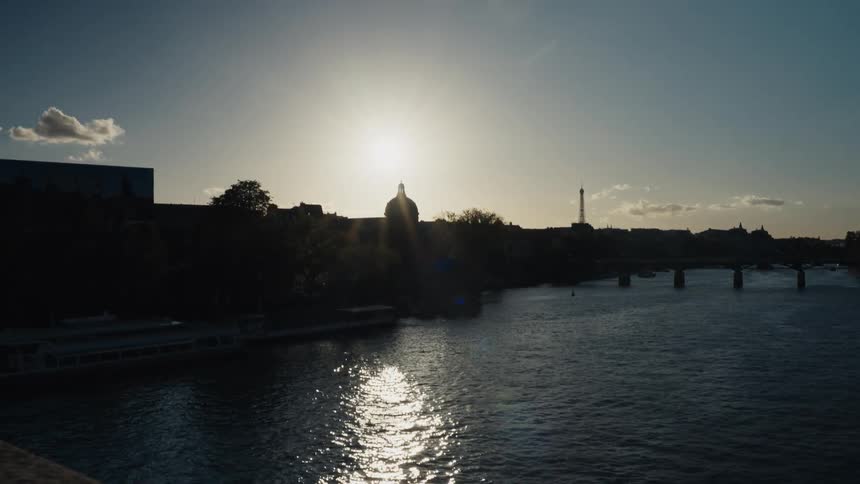 A serene sunset over the Seine River in Paris, with the city skyline and Eiffel Tower silhouetted against the bright sun as its light shimmers on the water