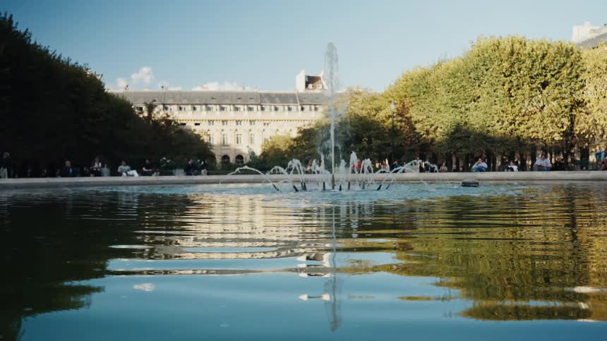 A tranquil water fountain reflects in a pond at a beautiful Parisian park on a sunny day
