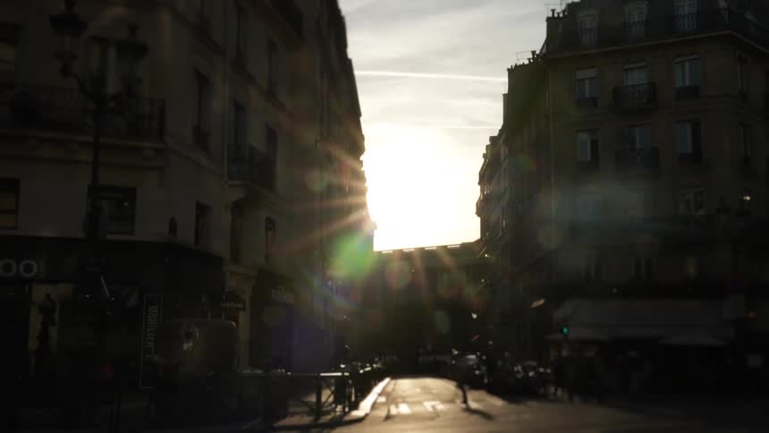 A dynamic time-lapse captures the vibrant energy of a Parisian street with cars and buses speeding by against a brilliant sunset flare