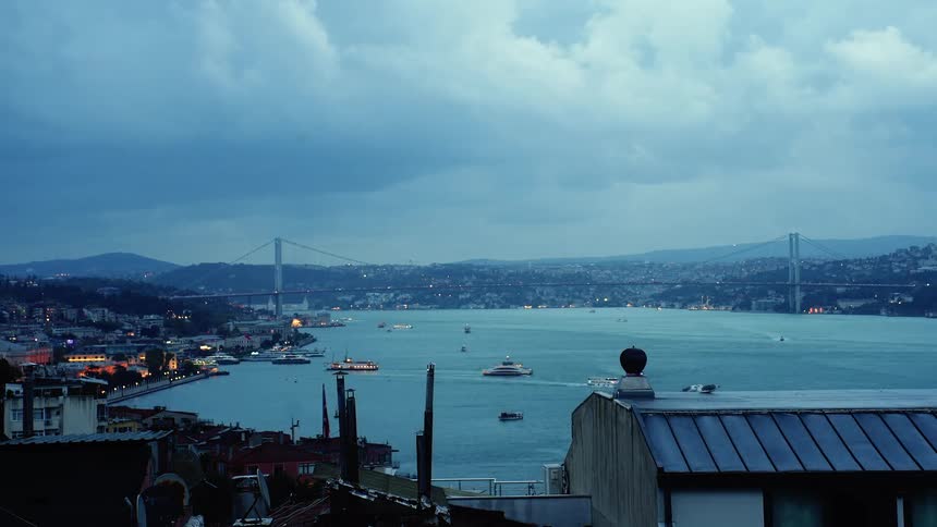 Dusk to night timelapse over Istanbul's Bosphorus Strait, showcasing the illuminated cityscape and busy boat traffic under cloudy skies