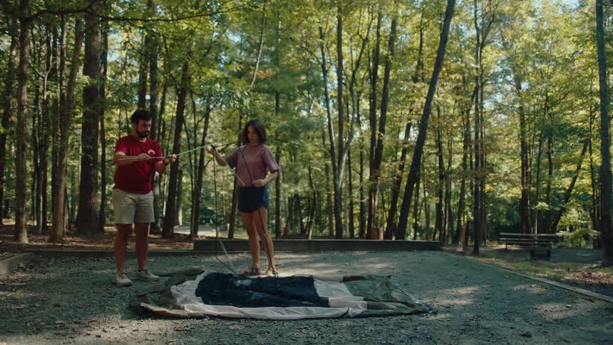 A young couple works together to assemble the poles and set up their tent at a campsite in the forest