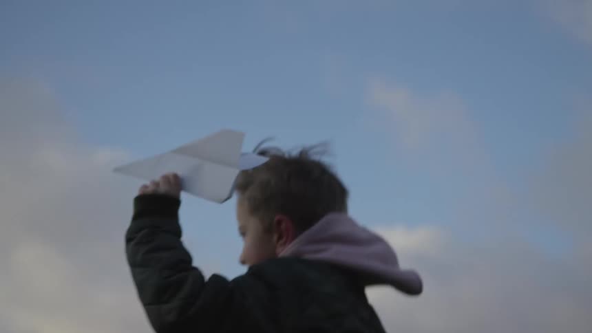 A young boy joyfully throws a paper airplane into the air against a cloudy sky in slow motion