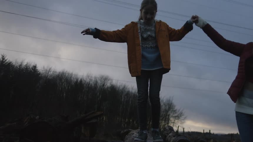 A young girl carefully balances on a log while holding her mother's hand for support in a clearing at dusk