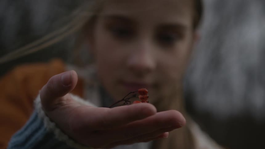 A young girl with windblown hair holds a sprig of red berries in the palm of her hand