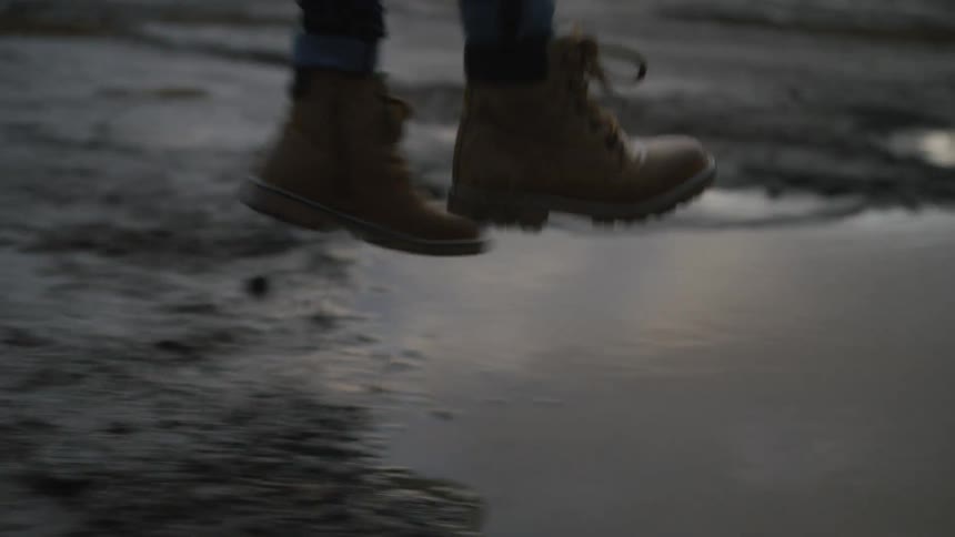 Slow motion close-up of a person's boots running and jumping into a muddy puddle, creating a dramatic splash