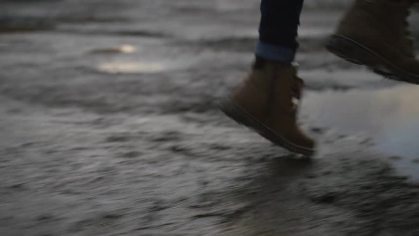 Slow motion shot of a person's feet in leather boots running and jumping into a large, muddy puddle