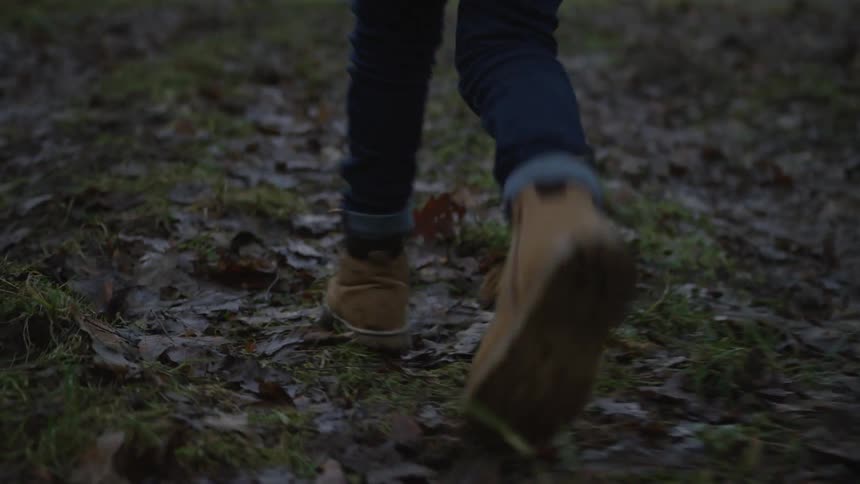A low-angle tracking shot follows a person's boots as they walk through a muddy, leaf-covered field during autumn