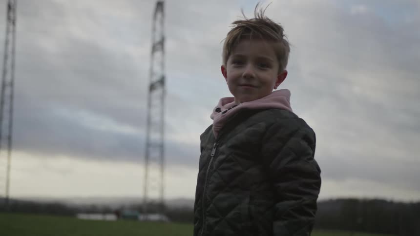A sweet young boy with windblown hair smiles at the camera in slow motion while standing in a field on a cloudy day