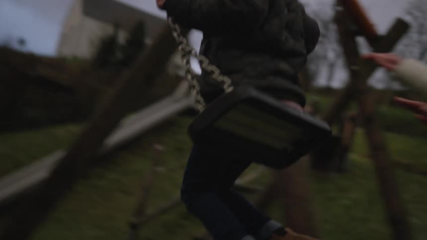 A mother lovingly pushes her young son on a swing at an outdoor playground on a gray, overcast day in slow motion