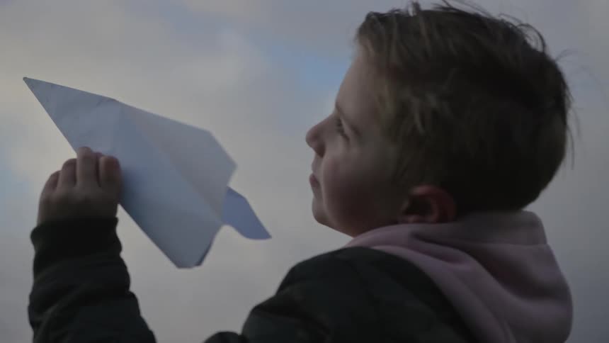 A young boy with a hopeful expression holds a paper airplane up to the overcast sky in a dreamy slow-motion close-up