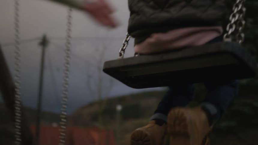 A mother gently pushes her young child on a swing set at the park in slow motion