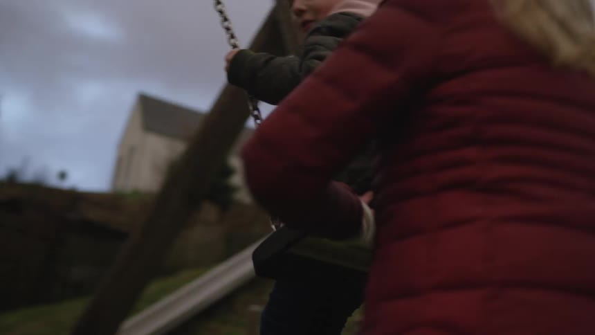 A mother gently pushes her happy son on a swing in slow motion at a quiet outdoor playground on a gloomy day