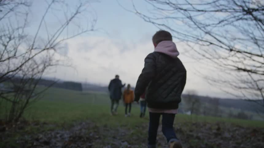 A young boy in a green jacket joyfully runs through a sparse forest to catch up with his family walking across a meadow in slow motion